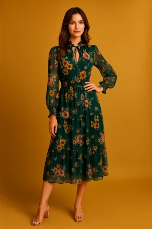 Woman in emerald-green floral midi dress with sheer long sleeves, ruffled tie neckline and tiered skirt, front view on a mustard backdrop.